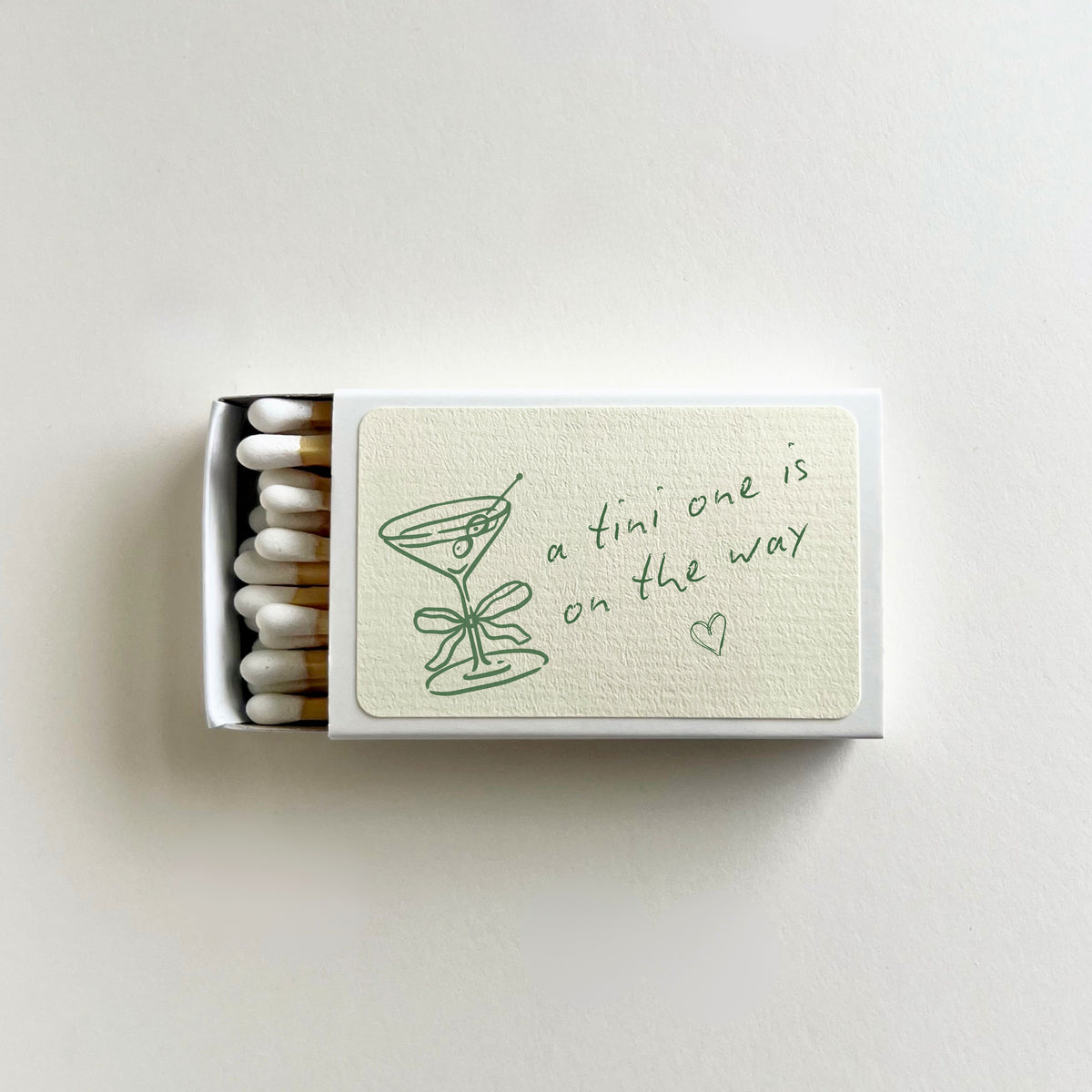 Personalized martini baby shower matchbook open to show white-tip matches, 'a tini one is on the way' text, and martini glass illustration