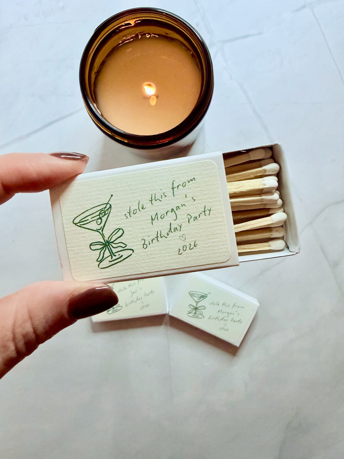 Hand holding open personalized martini birthday matchbox beside a lit candle, with two more matchboxes on a marble surface